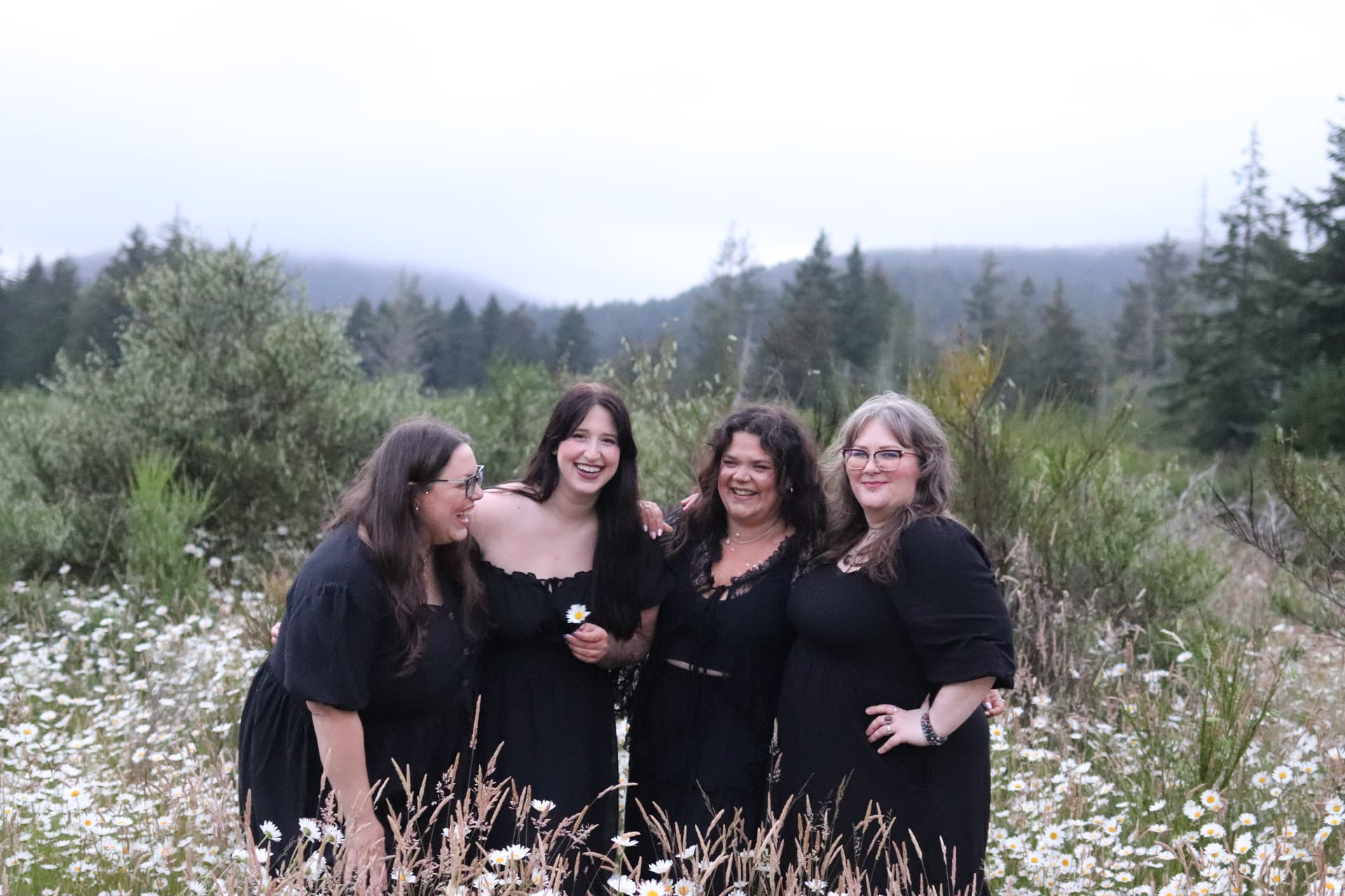 Four women in black dresses laughing together in a daisy field